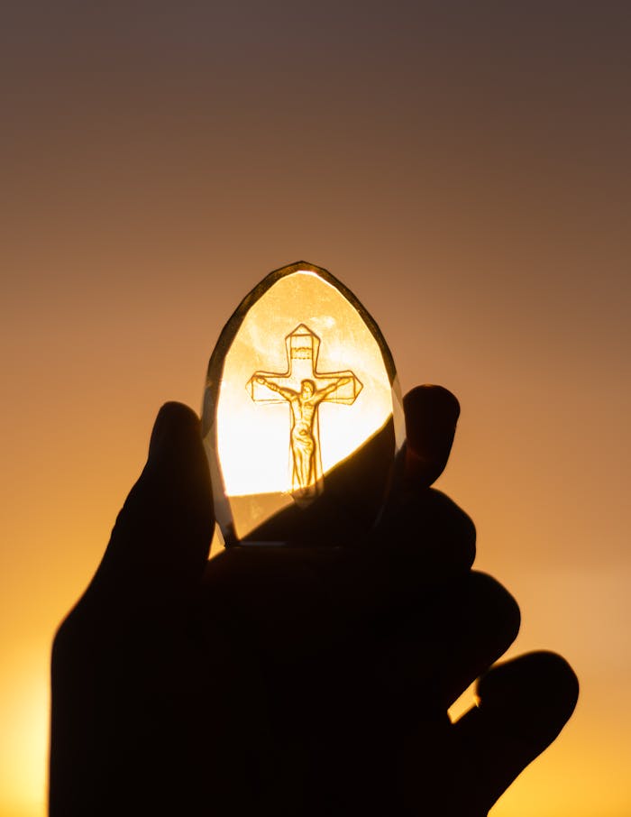 services-01 A silhouetted hand holds a crucifix against a vibrant sunset sky, symbolizing faith and spirituality.